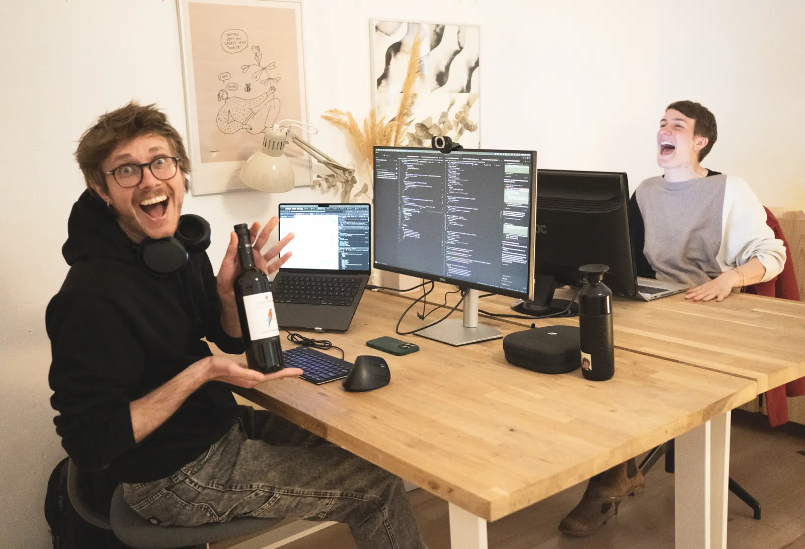 Zwei Menschen lachen an ihren Schreibtischen in die Kamera, einer hält eine Weinflasche hoch; auf dem Tisch Laptop und Bildschirme.