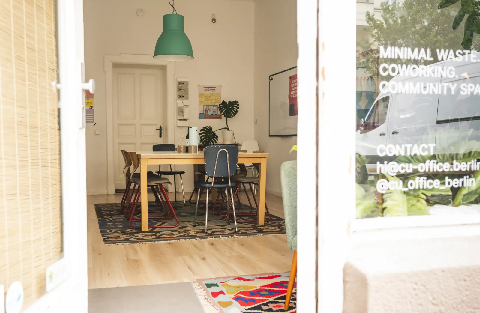 Blick durch die offene Ladentür in den Meetingraum mit Holztisch, Stühlen und grüner Hängelampe; auf der Scheibe steht Minimal Waste, Coworking, Community Space.