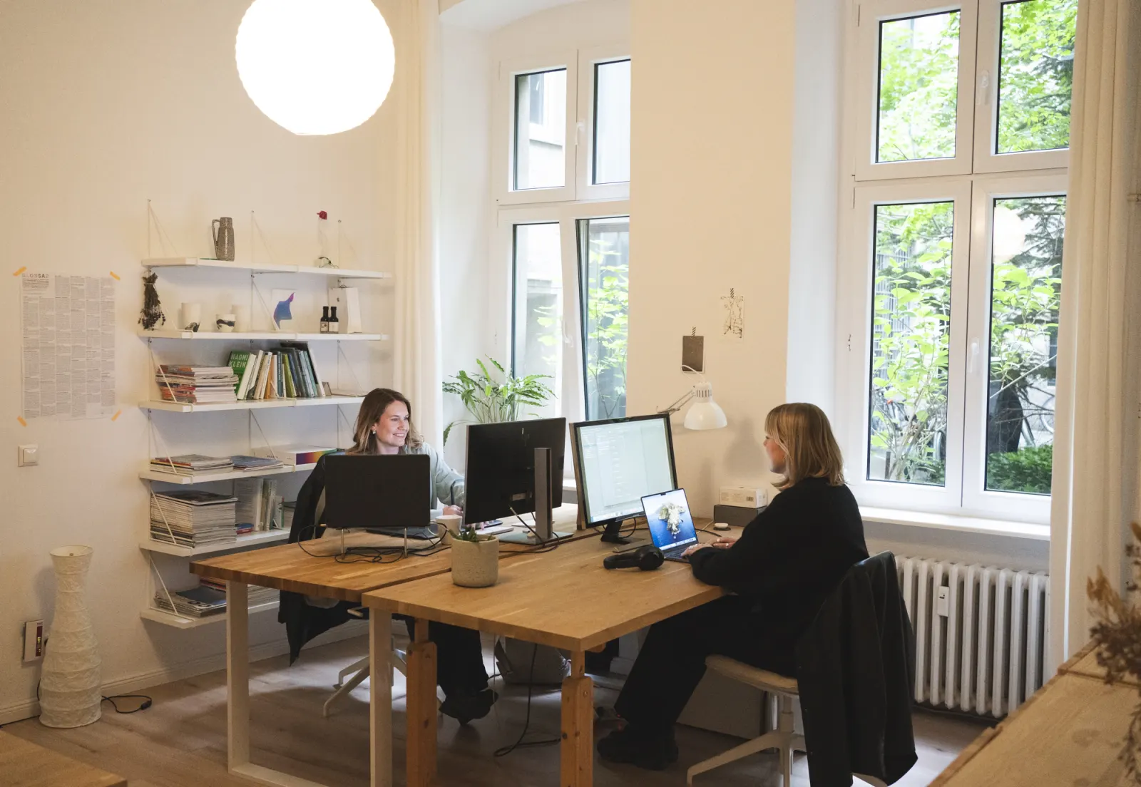 Zwei Frauen arbeiten an einem langen Holztisch mit Bildschirmen vor zwei hohen Fenstern, links ein Wandregal mit Büchern, darüber eine Kugellampe.