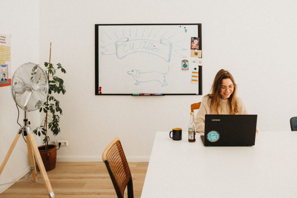 Eine Frau arbeitet lächelnd am Laptop an einem großen weißen Tisch, an der Wand ein Whiteboard mit dem gezeichneten CU.Office-Hund.