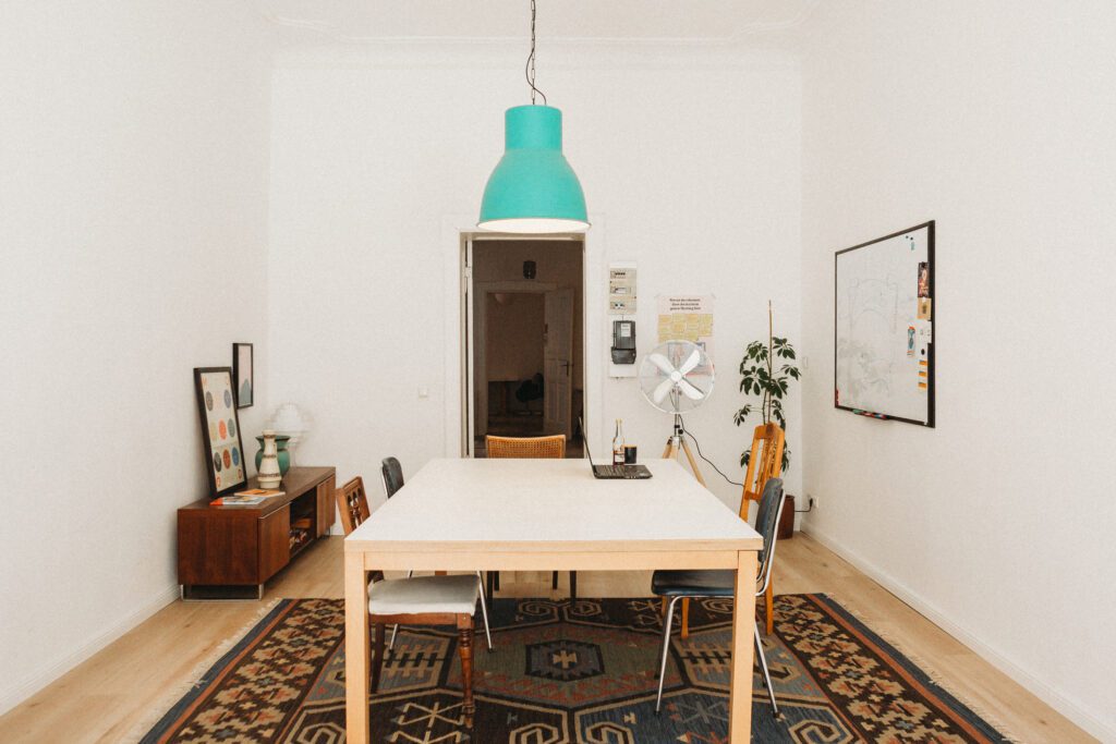 Meetingraum mit langem hellem Tisch, Stühlen, türkiser Hängelampe, gemustertem Teppich und Whiteboard an der Wand.
