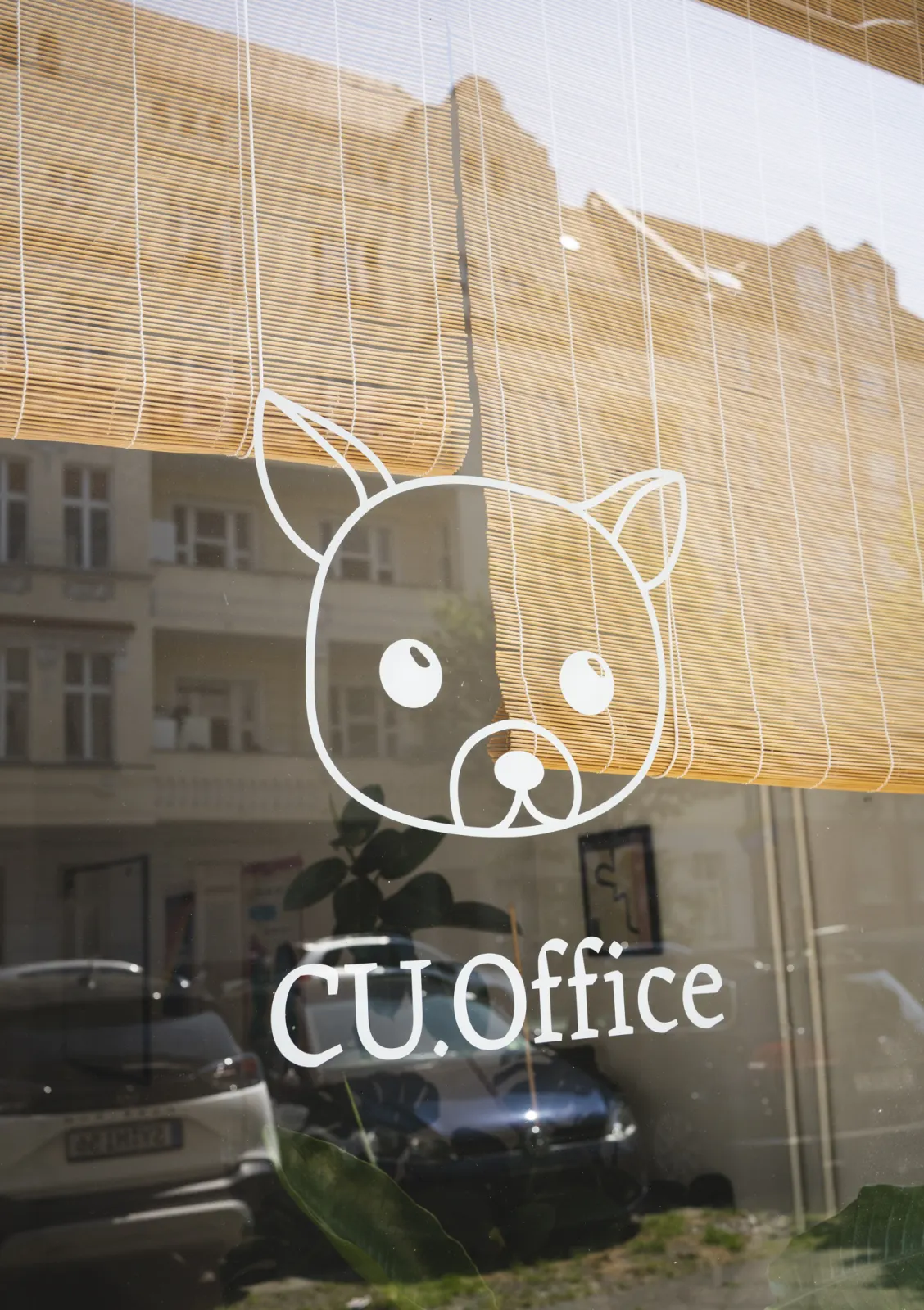 Schaufensterscheibe mit dem gezeichneten Hund und dem Schriftzug CU.Office, halb verdeckt von einem Bambusrollo; in der Scheibe spiegeln sich Altbauten und parkende Autos.