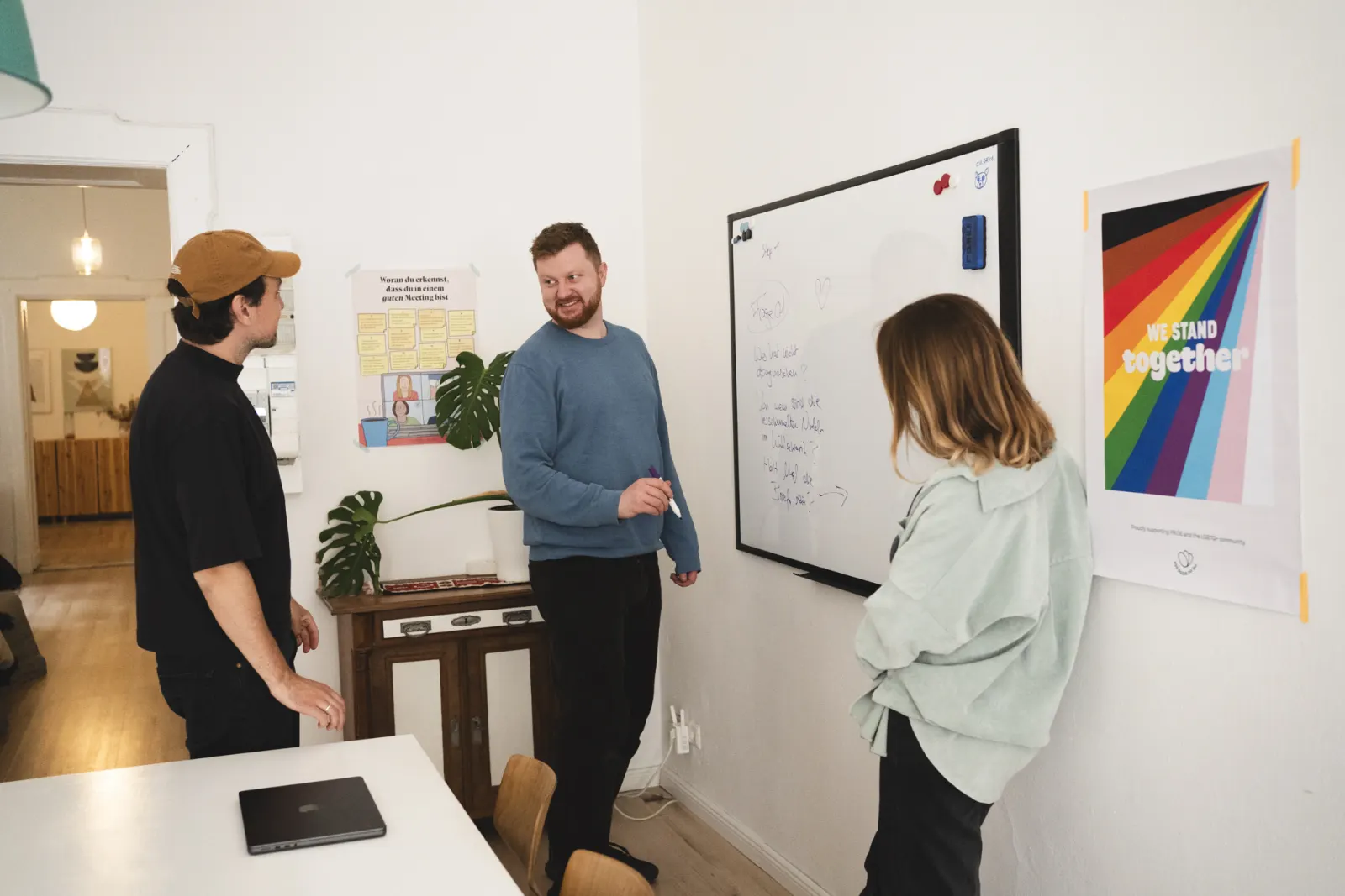 Drei Menschen stehen am Whiteboard und besprechen sich, an der Wand ein Regenbogen-Plakat mit dem Satz We stand together.